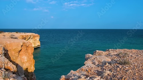 A scenic shot of the rugged coastline of Oman, featuring a dramatic view of rocky cliffs meeting the clear, turquoise waters of the sea under a bright blue sky. This footage captures the natural beaut