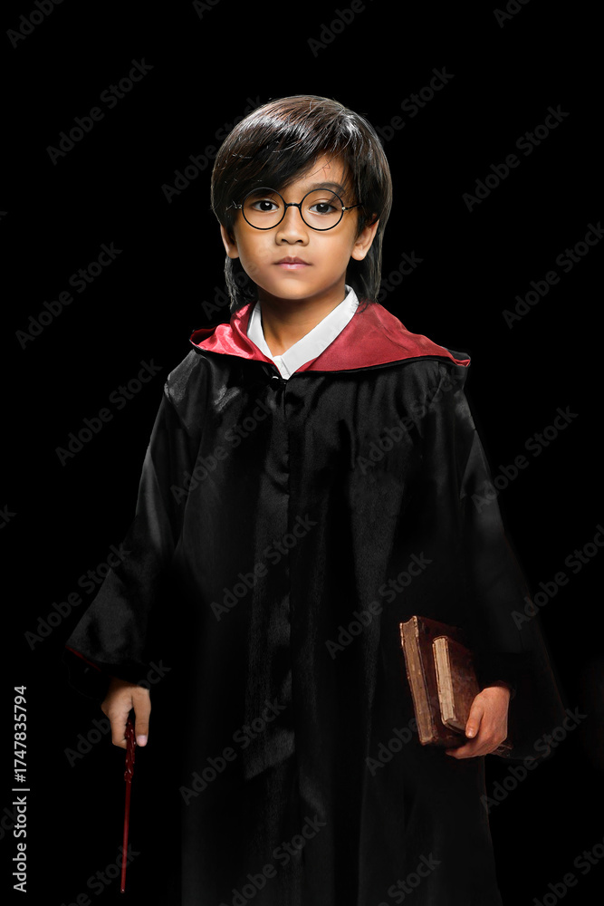 Fototapeta premium A young boy dressed as a wizard wearing black and red robe, holding a magic wand and old books while standing seriously against a black studio background. Fantasy, Halloween, or cosplay themes.