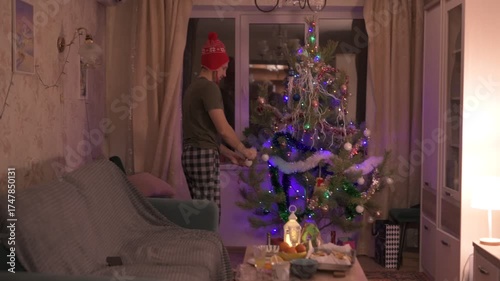 A young man in a red New Year's hat decorates a Christmas tree at home