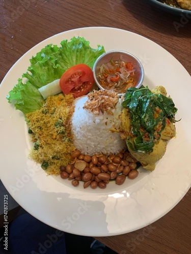 Balinese dish served on white plate featuring Bebek Betutu roast duck, accompanied by amound of rice topped with fried shallots. includes sauteed greens contain grated coconut, roasted peanuts, salad.