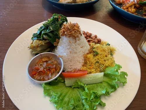Balinese dish served on white plate featuring Bebek Betutu roast duck, accompanied by amound of rice topped with fried shallots. includes sauteed greens contain grated coconut, roasted peanuts, salad.