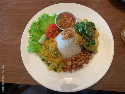 Balinese dish served on white plate featuring Bebek Betutu roast duck, accompanied by amound of rice topped with fried shallots. includes sauteed greens contain grated coconut, roasted peanuts, salad.