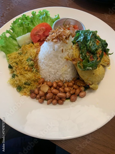 Balinese dish served on white plate featuring Bebek Betutu roast duck, accompanied by amound of rice topped with fried shallots. includes sauteed greens contain grated coconut, roasted peanuts, salad.