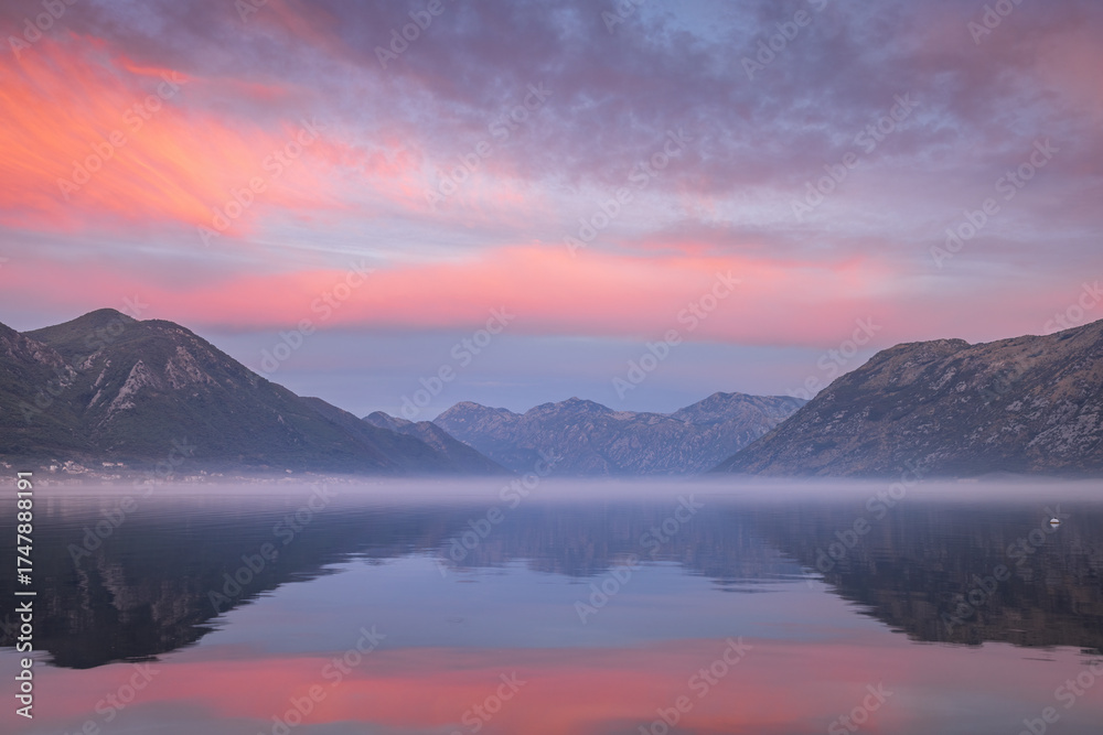 Obraz premium Misty dawn before sunrise over the calm Kotor Bay in Montenegro, with soft purple light, still water, and distant mountain silhouettes fading into morning fog.
