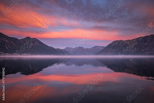 Misty dawn before sunrise over the calm Kotor Bay in Montenegro, with soft purple light, still water, and distant mountain silhouettes fading into morning fog.