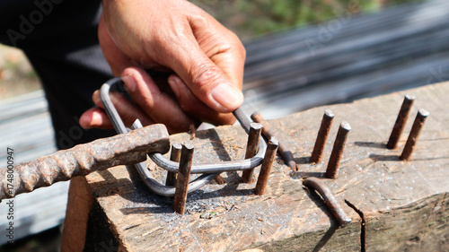 Worker manually bends steel rebar into stirrups using hammer and nail jig on wooden block. Ideal for construction, fabrication, and civil engineering process themes.