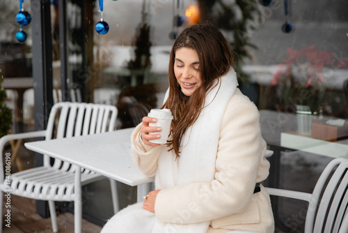 Smiling young woman in a white winter coat and scarf sitting at an outdoor café with a takeaway coffee. Snow and blue Christmas ornaments create a warm festive atmosphere.