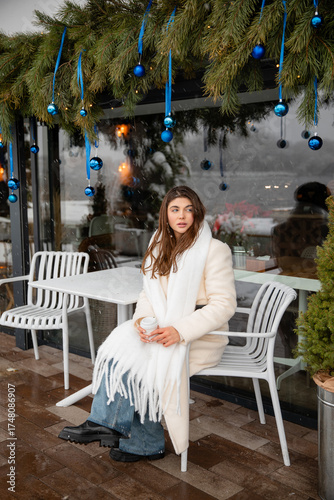 Smiling young woman in a white winter coat and scarf sitting at an outdoor café with a takeaway coffee. Snow and blue Christmas ornaments create a warm festive atmosphere.