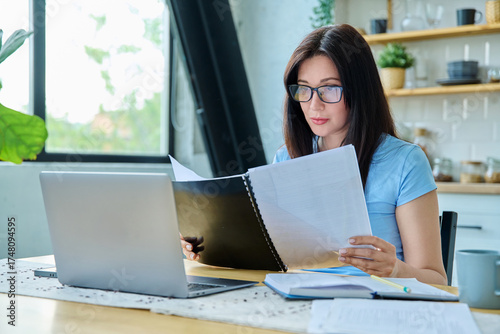 Young serious woman working with laptop, reading business papers, sitting at table at home