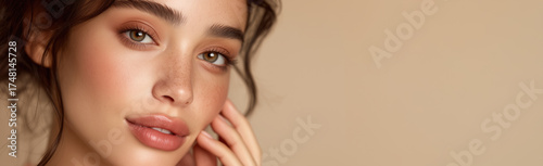 Woman with natural makeup and freckles touching face. Close-up beauty portrait on beige background with smooth skin and soft lighting
