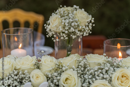 A beautiful gathering showcases a table adorned with white roses and baby’s breath, complemented by glowing candles. The setting exudes a tranquil evening ambiance.