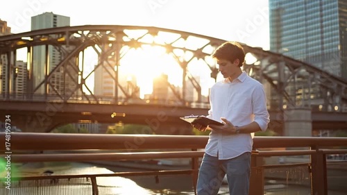 Young man reading clipboard by city bridge at sunset. Potential use Stock photo for business, education, or travel