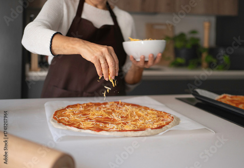 Obraz na plátne Woman's hands sprinkling shredded cheese over raw pizza dough with tomato sauce