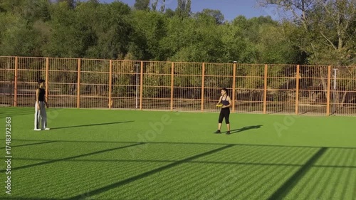Men in athletic wear tossing yellow frisbee disk across green artificial grass court, metal fence with trees visible behind, bright lighting.