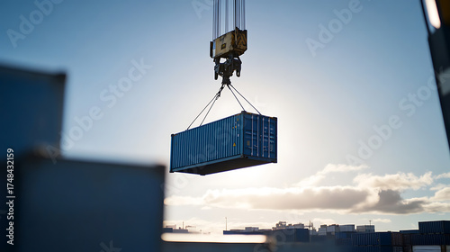 A cargo container is lifted by a crane against a bright, sunny sky. Other containers are visible in the background at a shipping terminal.