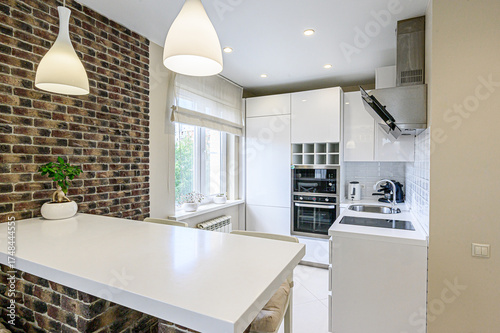 Bright kitchen with white cabinets, appliances, and countertops.  Features a brick wall accent and pendant lighting. The design is modern and minimalist