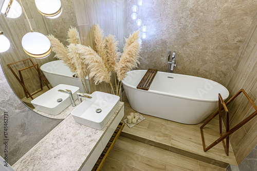 A stylish bathroom features a white bathtub, double sinks, pampas grass, and warm lighting. The aesthetic is modern with earthy tones and textures