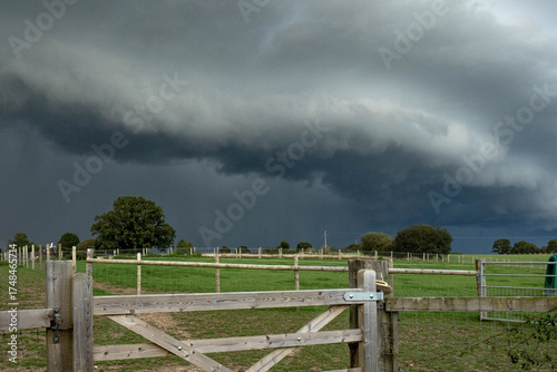 Dark storm clouds rolling across the English countryside, hovering above quiet green fields and wooden fences before a heavy summer rain.