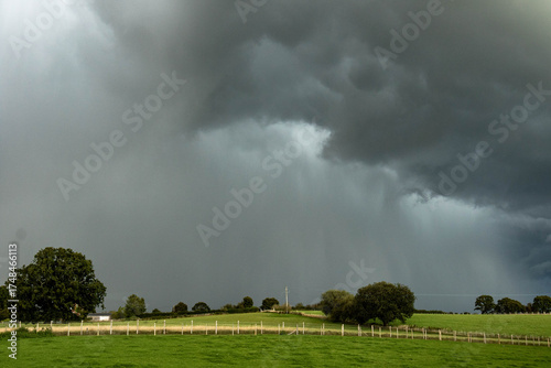 Dark storm clouds rolling across the English countryside, hovering above quiet green fields and wooden fences before a heavy summer rain.