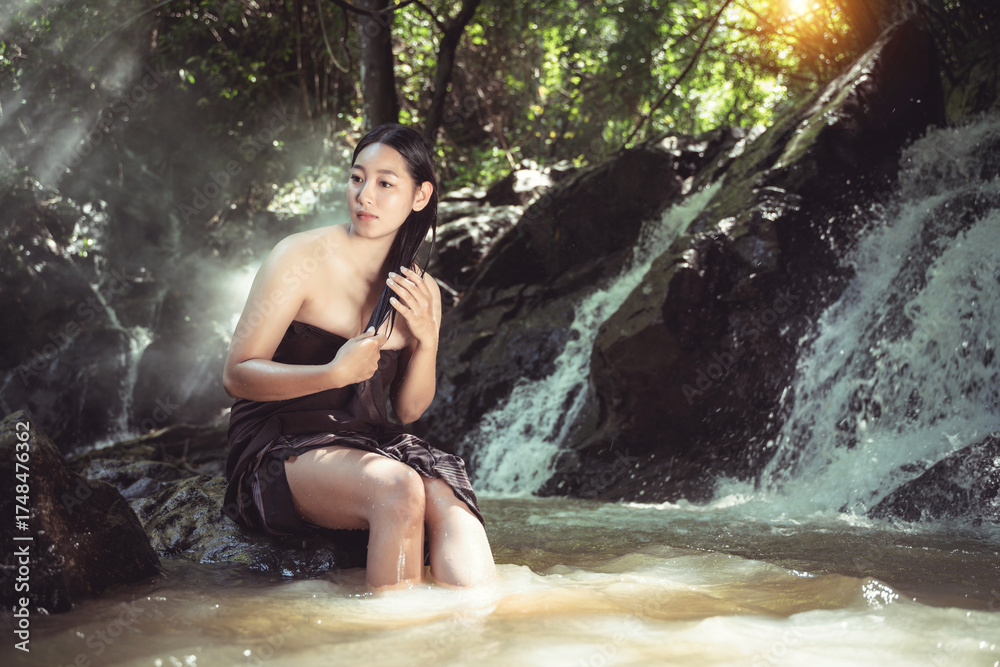 Fototapeta premium Serene Young Woman with Long Hair Sitting by Waterfall in Lush Green Forest, Soft Sunlight Filtering Through Trees, Peaceful Nature Scene