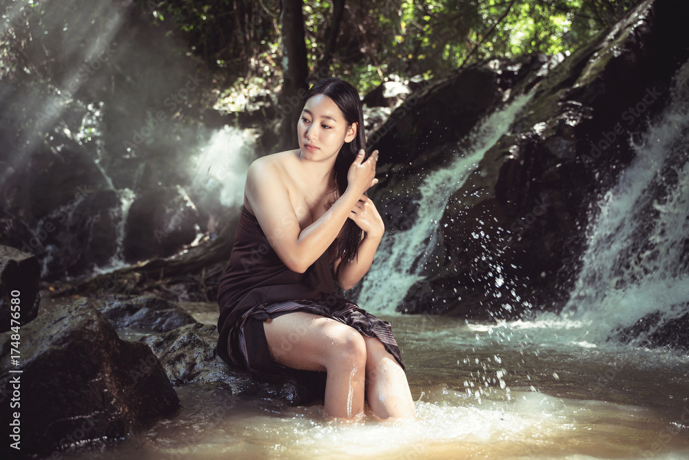 Fototapeta premium Serene Woman Sitting on Rocks in Waterfall, Natural Light Creating Dreamy Atmosphere, Refreshing Outdoor Scene with Lush Greenery and Calm Waters