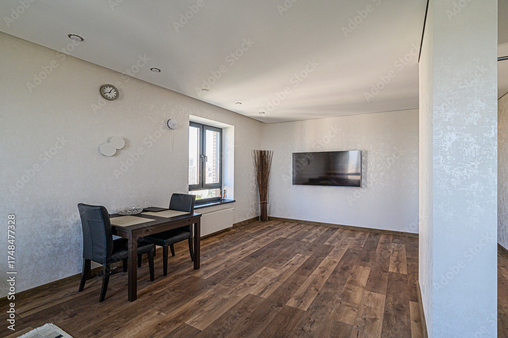Fototapeta premium modern living and dining area featuring dark wood flooring, a dining table with chairs, a wall-mounted TV, and natural light from a window. The decor is minimalist and clean