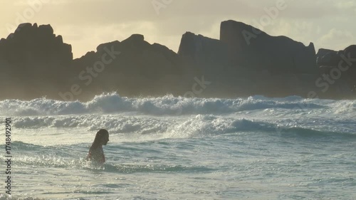 A woman enjoys a serene moment in the ocean during a golden Seychelles sunrise or sunset, flipping her hair in slow motion, with waves crashing around her in this stunning 4K vacation video.