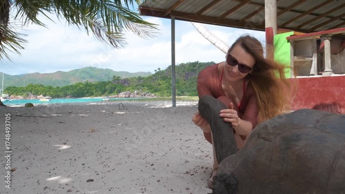 A smiling woman pets a giant tortoise on a tropical beach in Seychelles, with turquoise waters and green hills in the background. This 4K video captures a peaceful vacation moment.