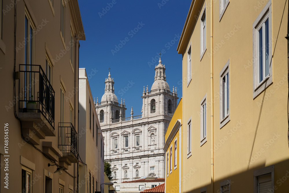 Fototapeta premium Street with residential façades leading to a historic structure with spires. Urban depth, architectural heritage, cultural layering, Portugal.