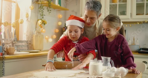 Grandmother teaching granddaughters how to roll dough while baking Christmas cookies together. Joyful family bonding in cozy kitchen filled with laughter and holiday spirit.