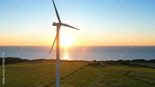Wind Turbine Dominating a Green Field against a Backdrop of Seascape at Sunset Under a Bright Sky with Golden Sunbeam