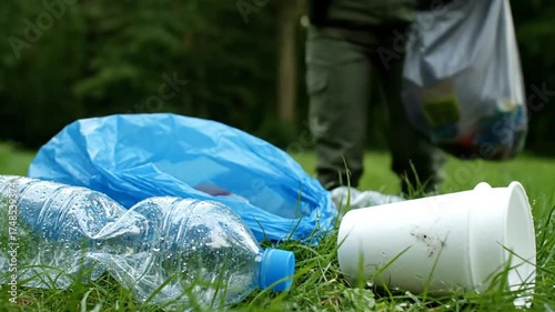 Close Up of Plastic Waste on Green Grass with Blurred Figure Holding Garbage Bag in Natural Outdoor Setting in Daytime Environment
