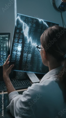 Vertical shot of female doctor seen from behind holding up and closely examining MRI brain scan when sitting at desk with computer equipment in front of her in clinical setting with dim lighting