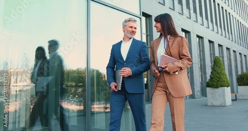 Adult man and young woman, dressed in formal business clothing, walk together toward the office while having work-related conversation, outside