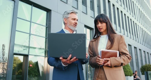 Confident Caucasian businessman in a suit holding a laptop, brainstorming with stylish businesswoman in a beige blazer outside a contemporary office building, outside