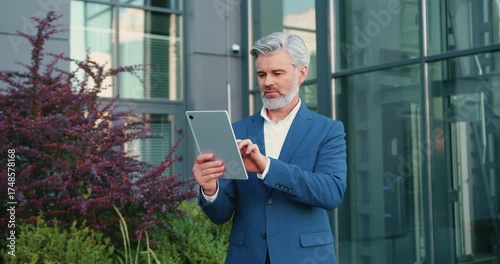 Confident adult businessman in suit working on tablet outside contemporary office building, focused on remote tasks in urban business environment