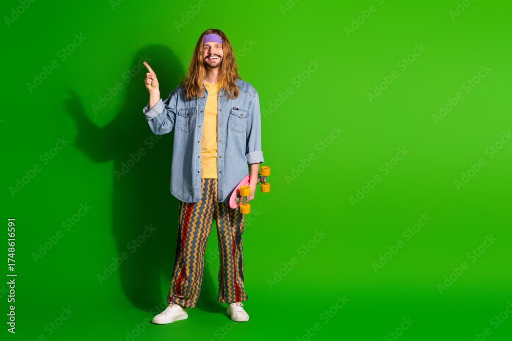 Fototapeta premium Young man with retro fashion holding skateboard and pointing up, standing against green background in vibrant casual style