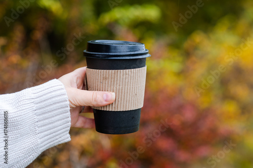 Female hand in white knitted sweater holding paper cup with hot drink on colorful autumn forest background