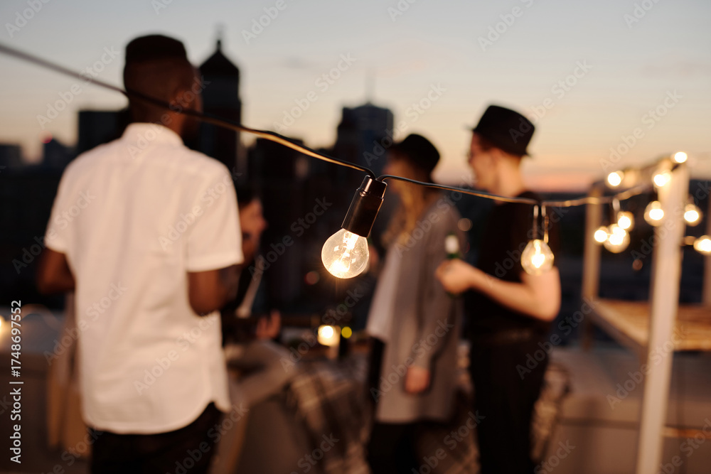 Fototapeta premium Close-up view of decorative lightbulbs illuminating rooftop where defocused young people standing and chatting on summer evening