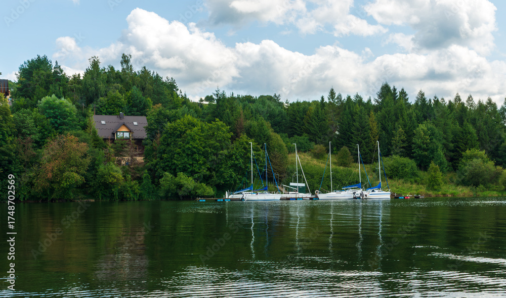 Obraz premium Peaceful sailboats rest at a small marina surrounded by lush green forest. The tranquil water reflects trees and white clouds, creating a serene summer atmosphere perfect for relaxation and nature tra