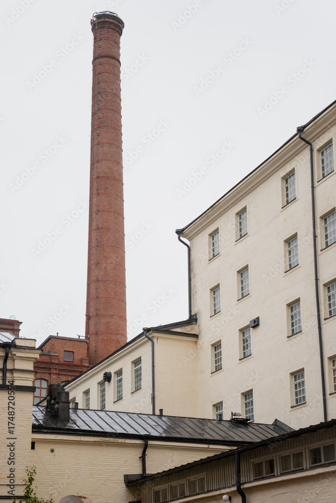 Fototapeta premium Historic brick industrial chimney and white plaster buildings in Tampere Finland representing preserved factory architecture, adaptive reuse design, and transformation of industrial heritage