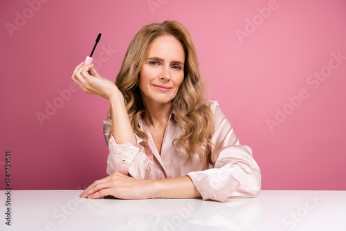 Beautiful mature woman with blonde hair holding mascara wand against pink background