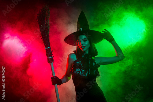 Young woman in witch costume holding broomstick surrounded by vivid green and red fog for a spooky Halloween theme
