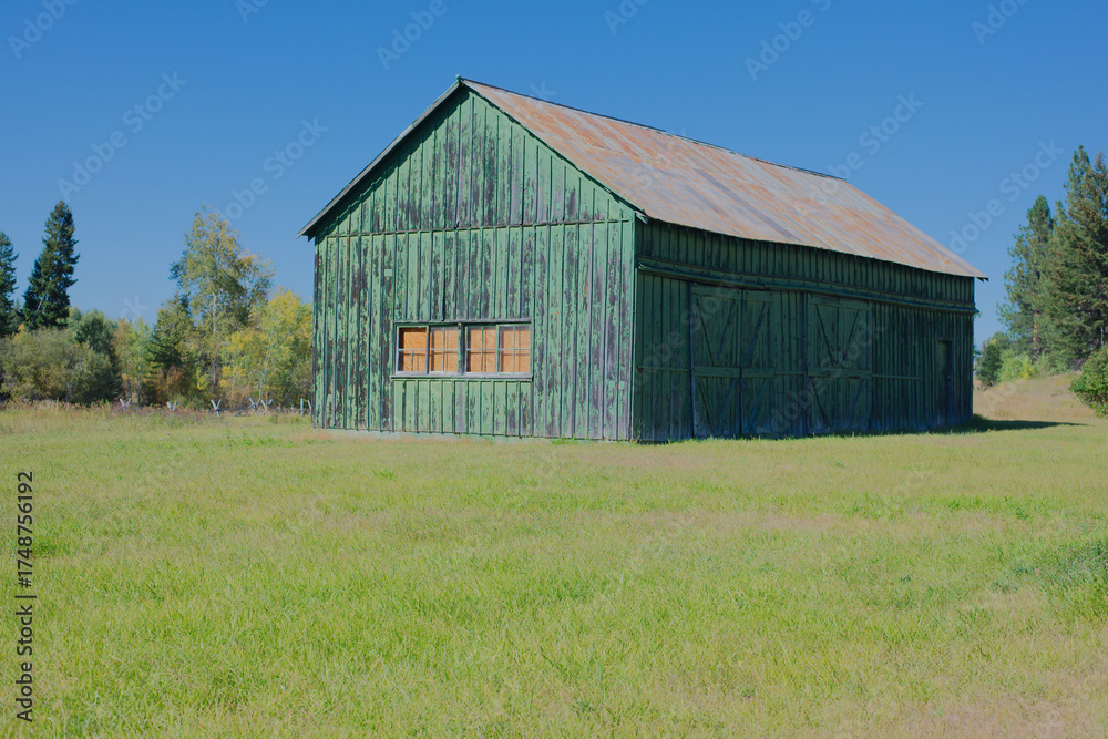 Obraz premium Rustic Wooden Barn With Weathered Logs On a Bright Green Field Under Clear Blue Sky. Historic Rustic Ant Flat Kootenai National Forest in Montana. Evoking countryside charm and rural life in a serene