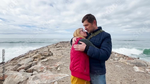 Happy middle-aged couple stands on a rocky breakwater by the Atlantic Ocean. They talk, kiss, and embrace lovingly, enjoying a romantic moment together under a cloudy sky near the sea