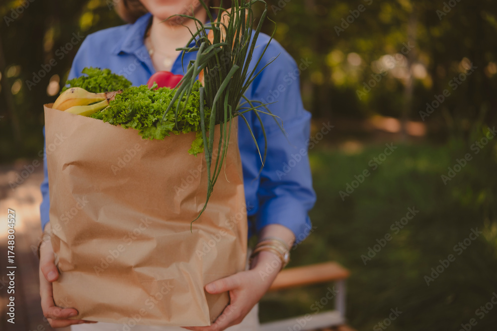 Obraz premium woman holds paper shopping bag with colorful vegetables, greens, and fruits in natural setting on sunny day