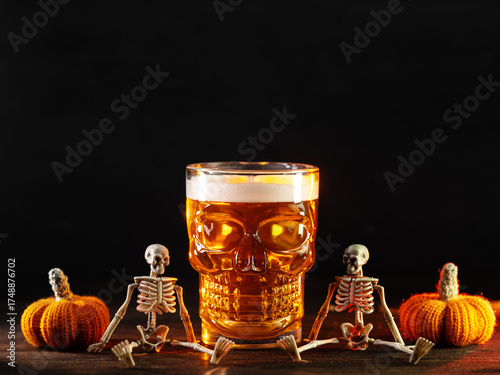 Spooky Halloween beer in a scull glass on a table with skeletons and pumpkins, copy space
