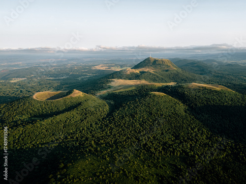 Aerial photo of Chaîne des Puys in Auvergne, France, showing volcanic craters, the Puy de Dôme summit, dense forests, and soft sunset light over a tranquil landscape.