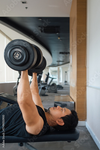 Asian male lifting dumbbells vertically on bench in gym