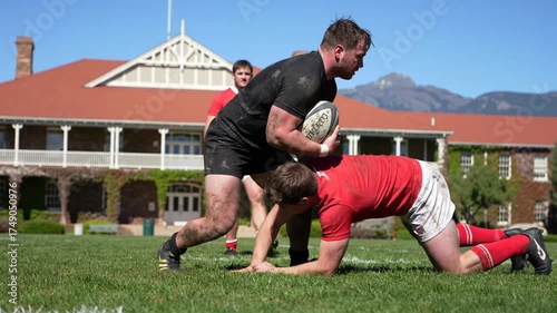 Dynamic low-angle wide video captures young adult male rugby players in black/red, intensely tackling on sun-drenched grass field, university/mountains in background, adrenaline-fueled sports